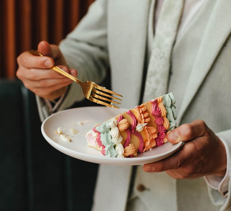 Groom in a pale green suit eating a colourful buttercream, retro wedding cake with a gold fork 