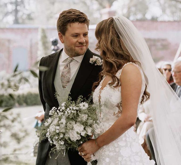 Groom looks intently into the bride's eyes as they stand at the altar together with groom in morning suit with green waistcoat and patterned tie 