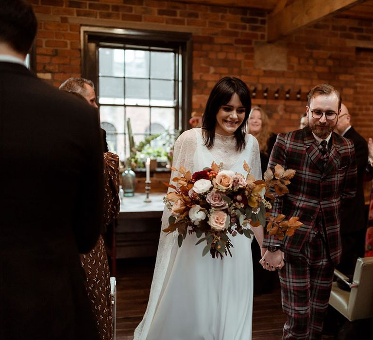 Bride & groom leave wedding ceremony hand in hand as bride holds Autumnal floral bouquet and wears lace cape 