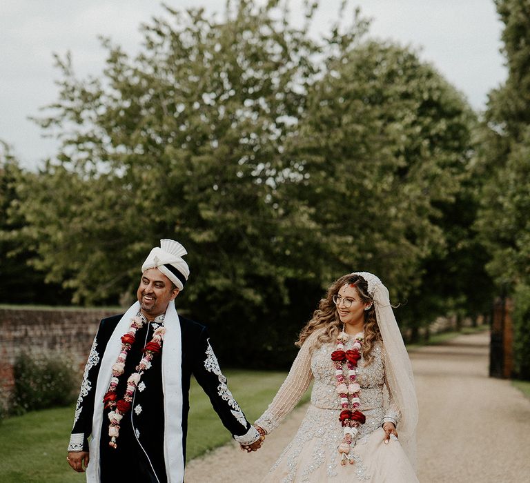 Bride & groom walk hand in hand outdoors wearing traditional garlands around their necks 