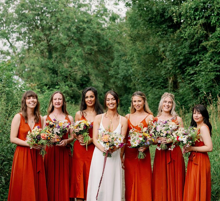 Bride stands with her bridesmaids who wear orange silk slip dresses 