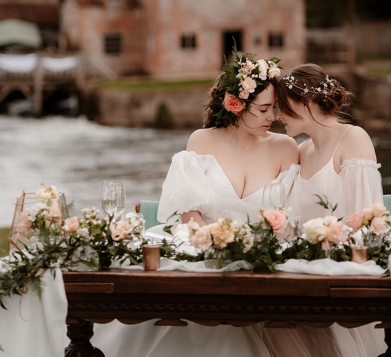 Intimate portrait of a bride in an ethereal wedding dress with sheer sleeves with her bride in a strapless wedding dress and flower crown at their outdoor sweetheart table 