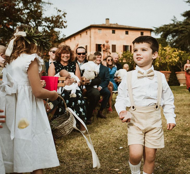 Page boy walks down the aisle wearing white shirt and sand coloured bow tie with braces and shorts 