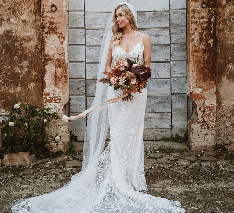 Bride stands in front of rustic wall whilst holding Autumnal floral bouquet and wearing romantic lace wedding dress with low cut front