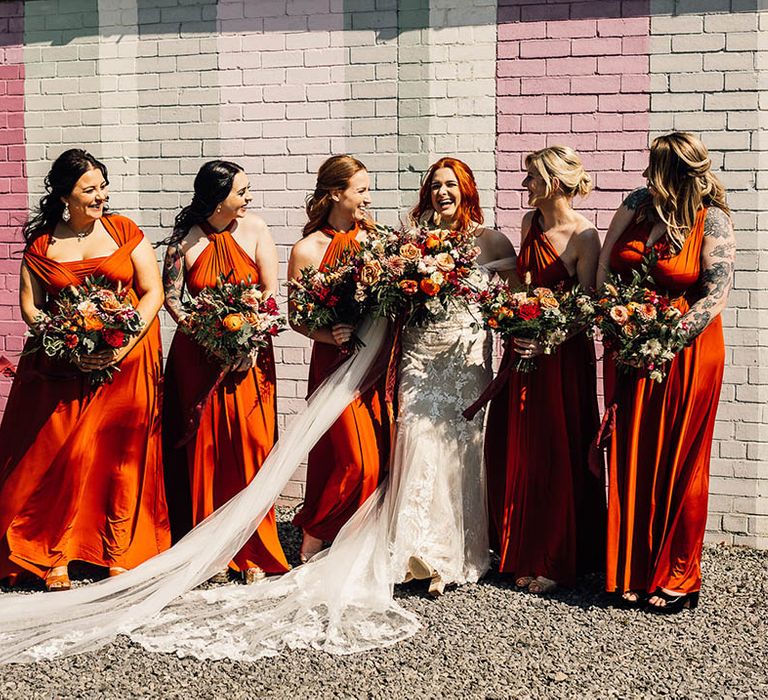 Bride laughs with her bridesmaids in orange dresses with matching autumnal bouquets in front of a colourful painted wall 