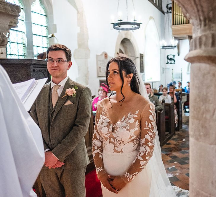 Bride wears sheer lace embellished wedding gown complete with floor-length veil and tendrils of her hair framing her face 