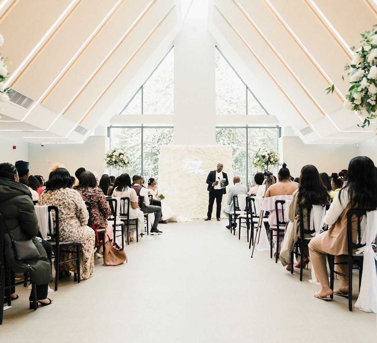 The Edgbaston Park Hotel wedding ceremony room complete with large floral arrangements to the sides of the aisle and statement white rose flower wall with neon sign