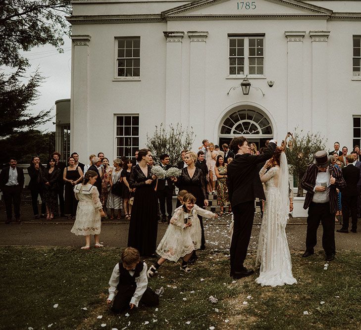 Bride & groom stand outside Belair House after wedding ceremony as white confetti surrounds them and bridesmaids wear black bridesmaid dresses