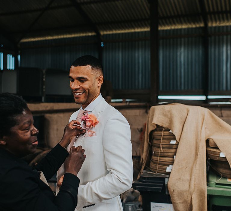 Groom in a white suit and pink tie gets his pink flower buttonhole attached 