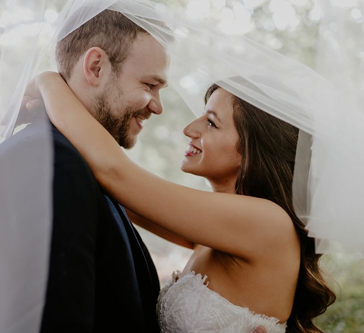 Bride & groom stand beneath veil as bride wears strapless lace wedding dress 