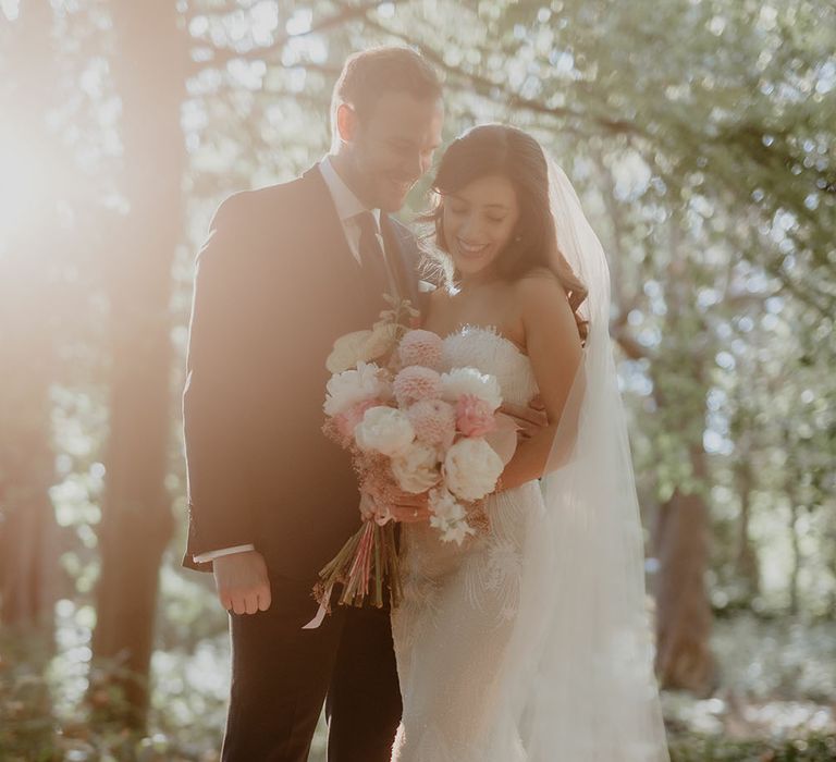 Bride wears fitted lace Pronovias wedding dress and floor-length veil and holds white and pink floral bouquet whilst stood beside her groom during golden hour