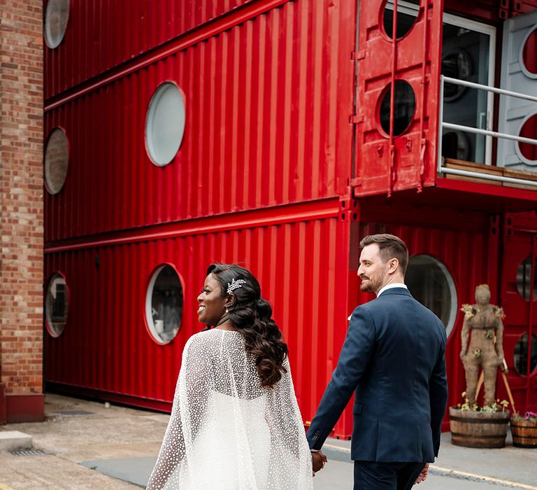 Bride wears tulle wedding cape with glitter polka dots and walks alongside her groom in front of bright red background 