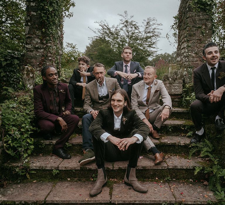 Groom in green suit sits with wedding guests on staircase outdoors for Autumnal boho festival styled wedding