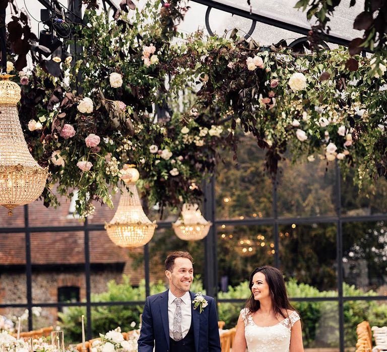 Bride in Phillipa Lepley applqiey wedding dress with groom in three piece blue suit walking in their glass marquee reception with chandelier installation