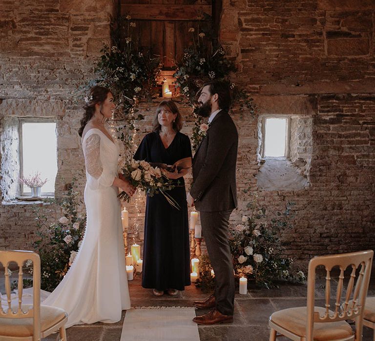 Bride wearing a sheer long sleeve wedding dress with the groom in a chocolate brown suit standing at the altar together with flower column decor 