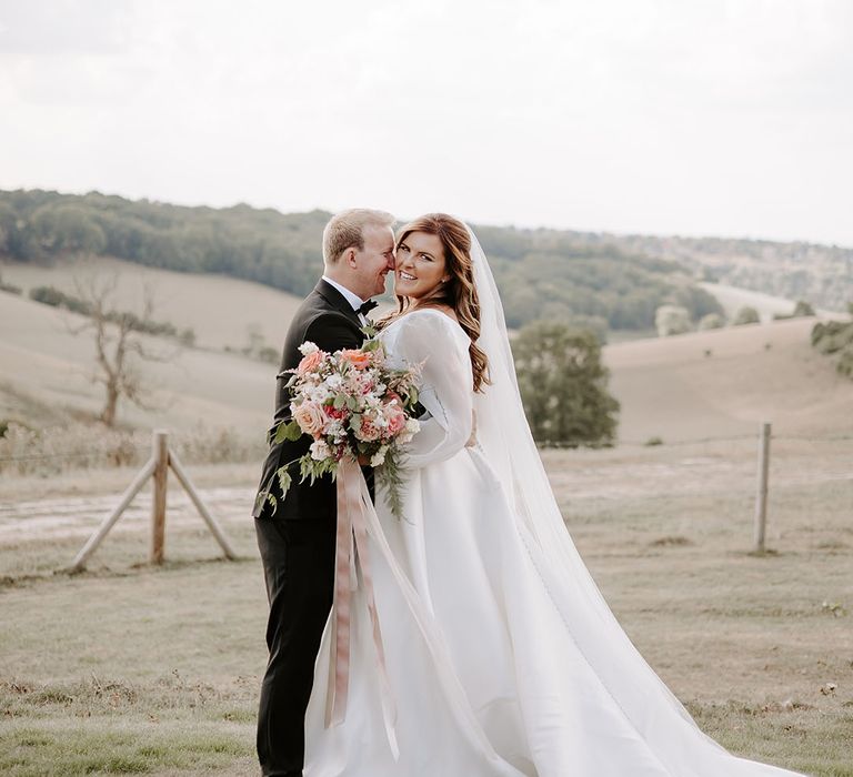 Bride holds colourful floral bouquet tied with silk pink ribbon stands with her groom outdoors 