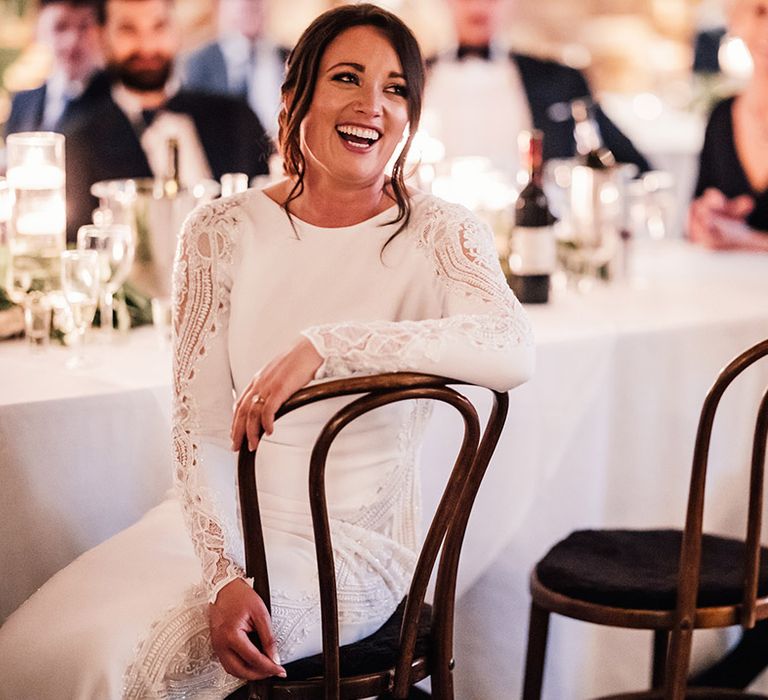 Bride in a long sleeve fitted wedding dress sits laughing during the wedding speeches 