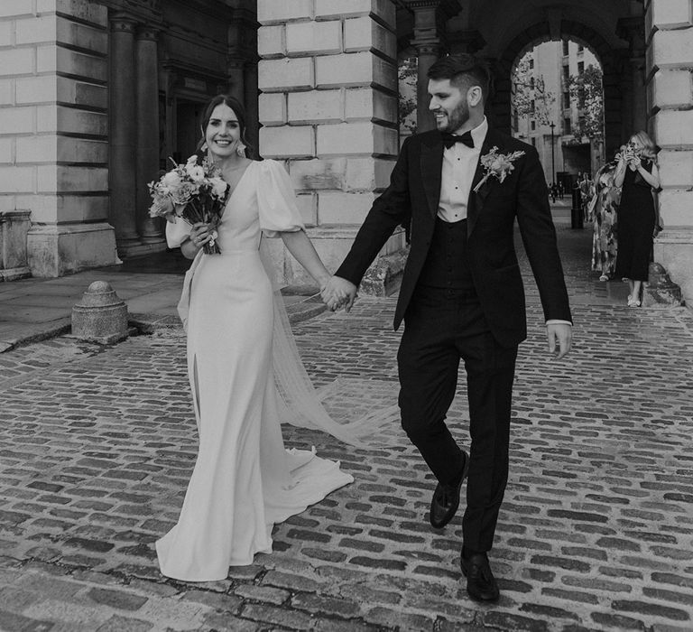 Bride in stylish modern wedding dress with puff sleeves and front slit holding hands of groom in dark tux