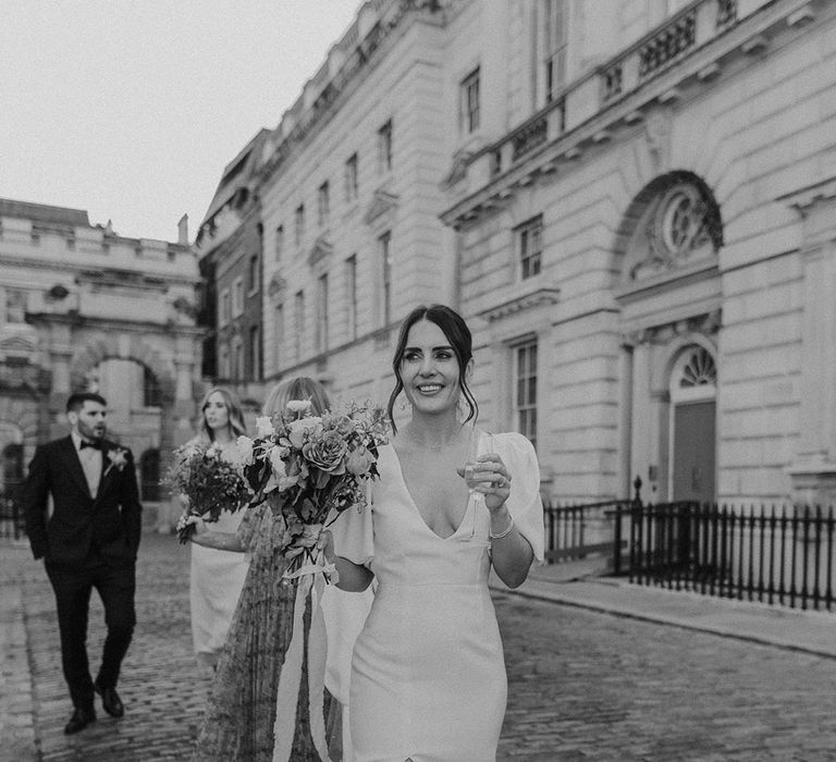 Bride in stylish modern wedding dress with puff sleeves and front slit holding bouquet and champagne glass