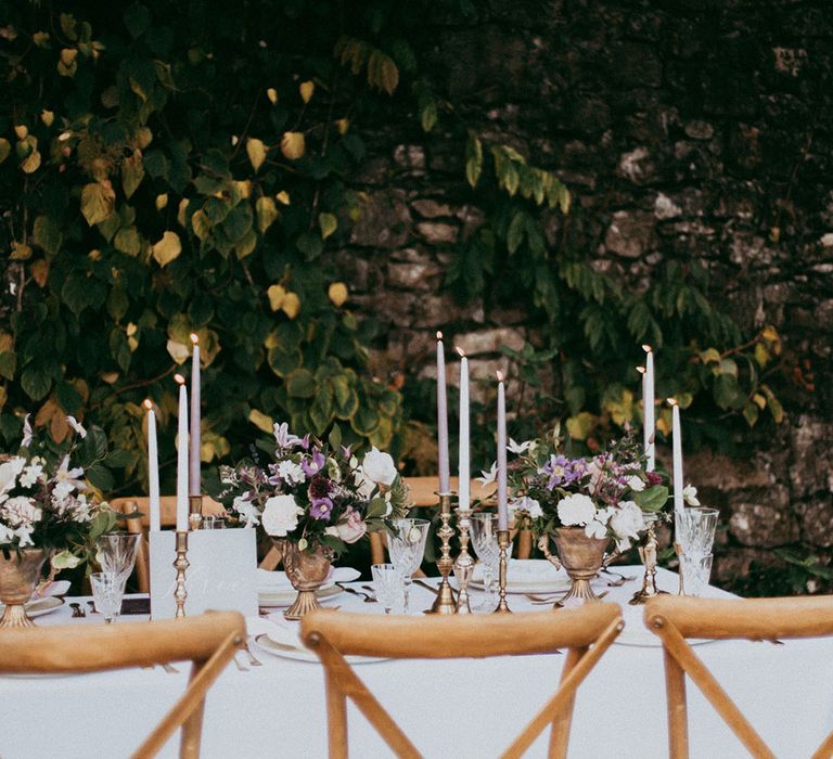 Wedding tablescape with lavender and white tapered candles in gold candle holders and mixed neutral flower arrangements with white carnations, purple tulips, roses and foliage 