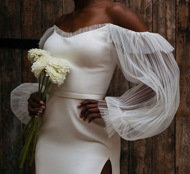 Bride in off shoulder wedding dress with front slit and dramatic lace puff sleeves holding bunch of white dahlias at 100 Barrington