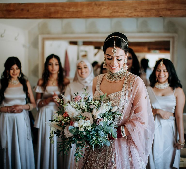 Bride wears golden bridal Lehnga while holding a bridal bouquet consisting of champagne coloured flowers, bunny ears and eucalyptus. Her bridesmaids are behind her wearing champagne bridesmaid dresses and bridesmaid separates