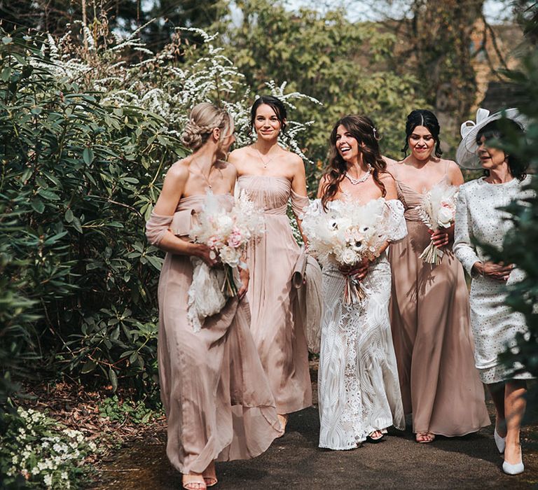 Bridesmaids in neutral pink dresses walking with the bride in her lace gown and the mother of the bride in a matching white dress and hat