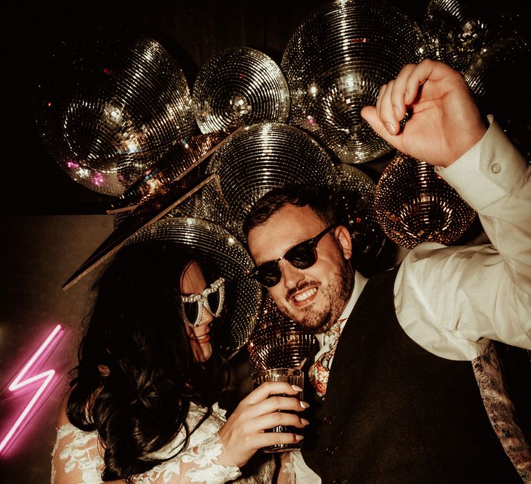  Groom in black waistcoat and flower patterned tie wearing Raybans sunglasses and bride in off the shoulder bridal gown with long, sheer sleeve and lace detailing wearing cat eye bejewelled sunglasses dancing by large disco ball wedding decor and neon signs