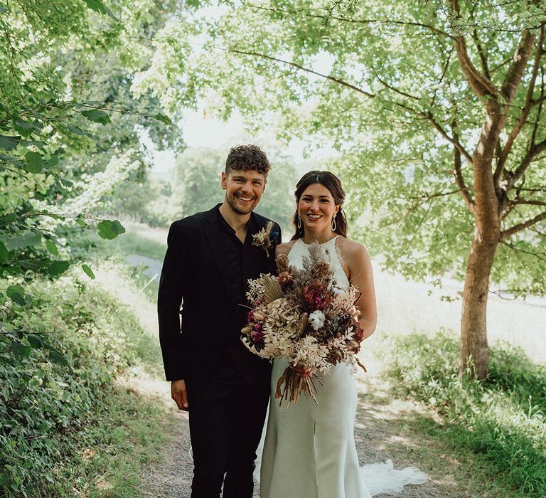 Bride in a halter neck wedding dress with front slit holding neutral wedding bouquet smiling with the groom in an all black suit 