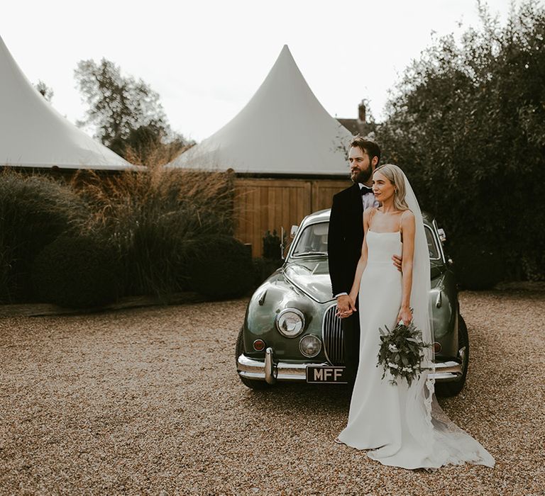 Groom in black tie embracing the bride in a fitted simple wedding dress in front of their green vintage wedding car 