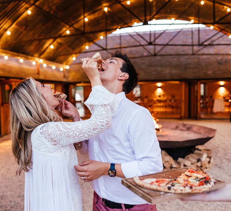 Bride in boho long sleeve wedding dress feeding pizza to groom in white shirt, black belt and pink corduroy trousers