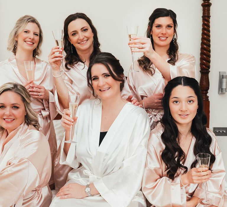 Bridesmaids in satin pink robes drink a glass of champagne with the bride in a white satin robe as they get ready for the day 