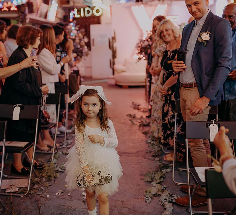 Flower girl wearing large bow, tutu and lace top sprinkling flower petals down the aisle 