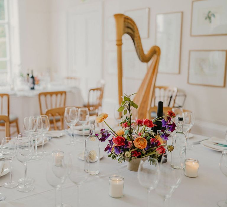 Gold harp beside round tables covered with white tablecloth finished with brightly coloured floral centrepiece 