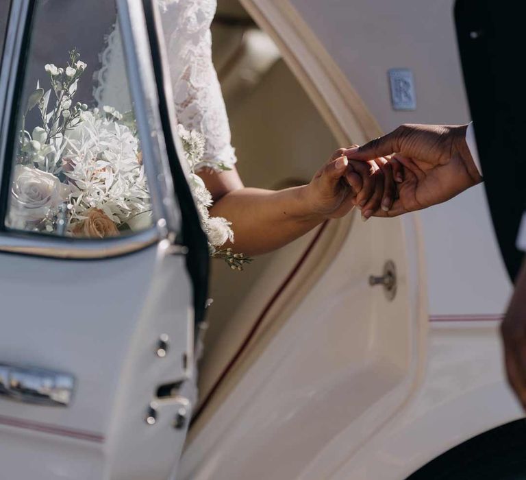 Bride in lace wedding dress holding large mixed floral bouquet getting out of classic white wedding car 