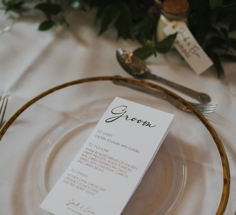 Black and white wedding menu on the clear charger plates with gold rim on white tablecloth with foliage table runner