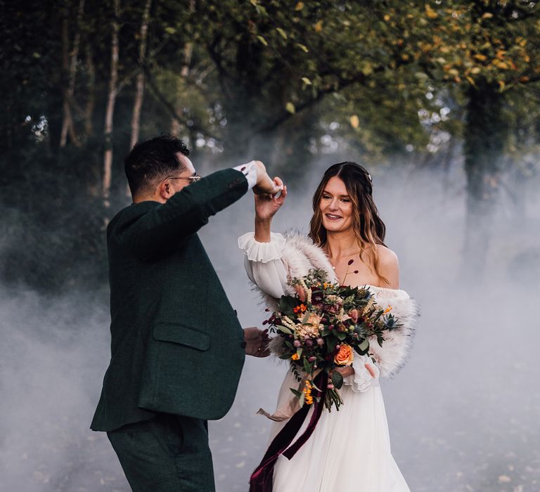 Bride in off the shoulder long sleeve wedding dress with winter faux fur shawl dancing with the groom with smoke bomb