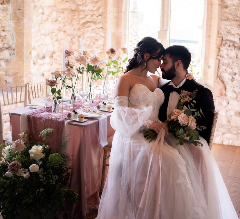 Groom in classic black tuxedo with bowtie and garden rose and eucalyptus boutonniere sitting with bride in ethereal off the shoulder wedding dress with detachable puff tulle sleeves and embroidered rose 3d appliqué details 