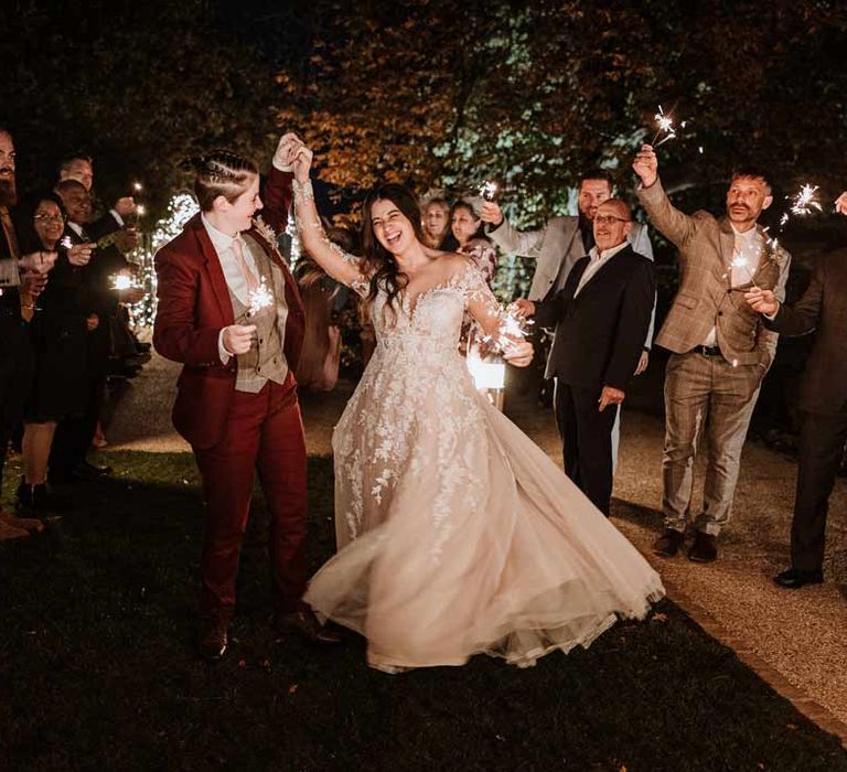 Bride in maroon bridal suit with grey waistcoat, dusky pink tie and autumnal dried flower boutonniere dancing with bride in wearing long sleeve lace illusion sleeve wedding dress with overlay during sparkler exit 