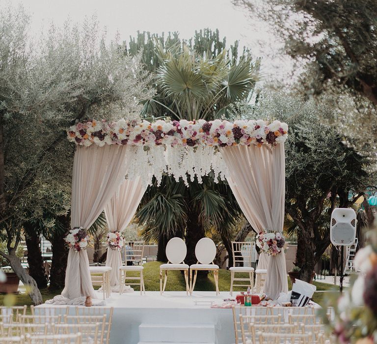 Traditional Indian wedding mandap with light blush fabric, garden rose, peony, eucalyptus and dried flower garlands and white chairs at the end of the wedding aisle at Barcelona fusion wedding 