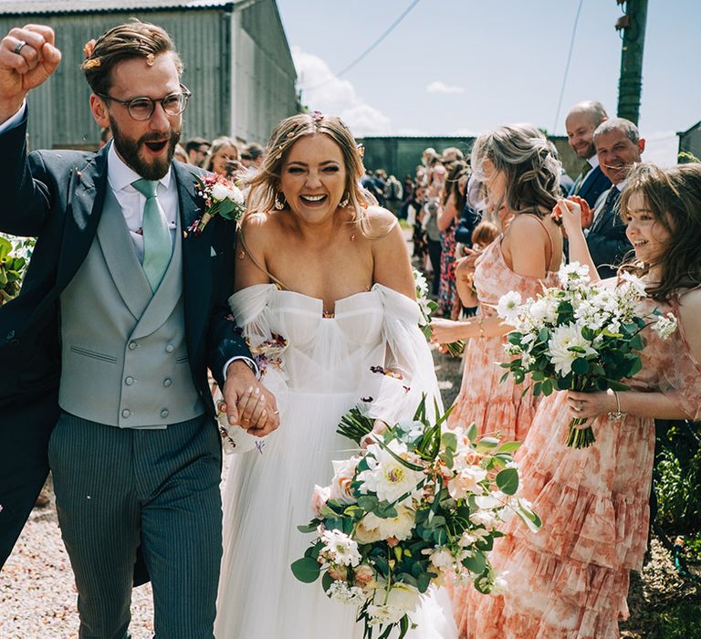 Groom wearing blue and grey double breasted morning suit celebrating with the bride in tulle wedding dress as they walk out to confetti 