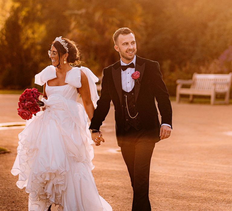 Groom in black tie walking with the bride in a ruffle wedding dress with bow straps holding pink rose bouquet 