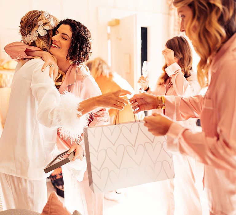 The bride in white satin pyjamas hugs the bridesmaids in pink satin pyjamas while they exchange gifts 