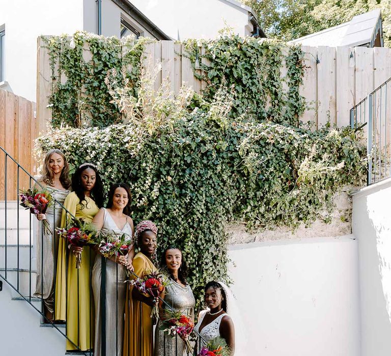 Bride in v neck sleeveless lace Pronovias wedding dress, cathedral length veil and silver sparkly bridal jewellery holding colourful tropical African bridal bouquet standing with bridesmaids in yellow, silver and gold mismatched bridesmaid dresses