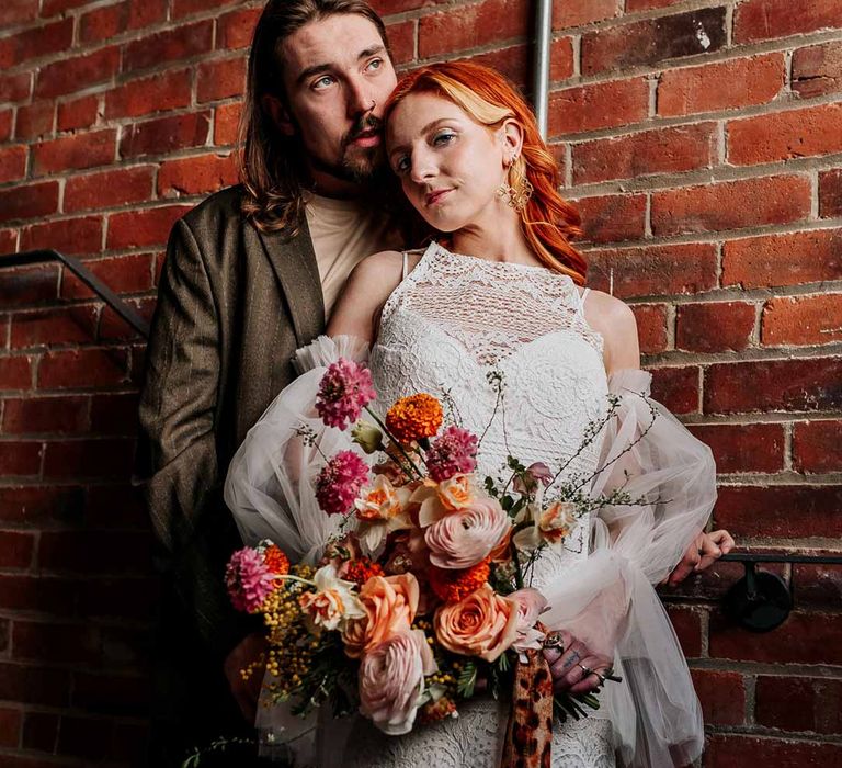 Bride in 70s-inspired boatneck short lace wedding dress with detachable tulle puff sleeves holding large colourful mixed flower bridal bouquet with groom in brown blazer, white shirt, grey suit trousers and vans shoes 