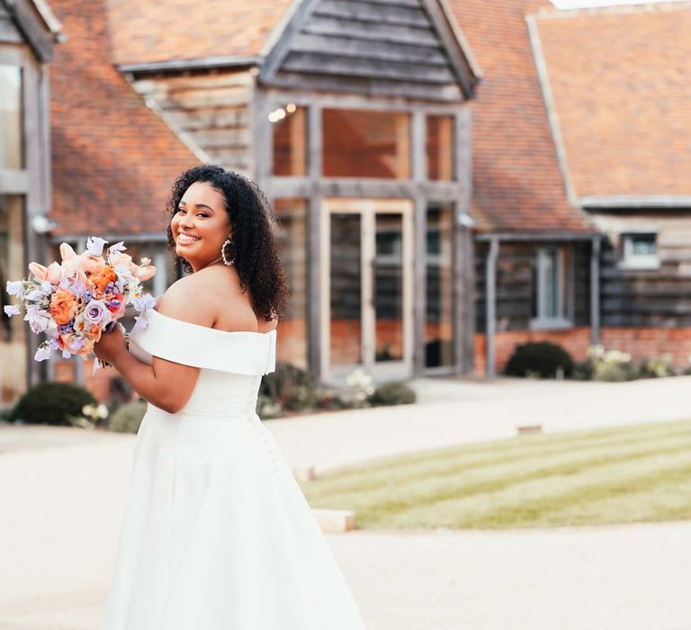 Bride in off the shoulder wedding dress with puddle train holding orange, lilac and peach mixed flower bridal bouquet standing outside Rackleys Barn wedding venue