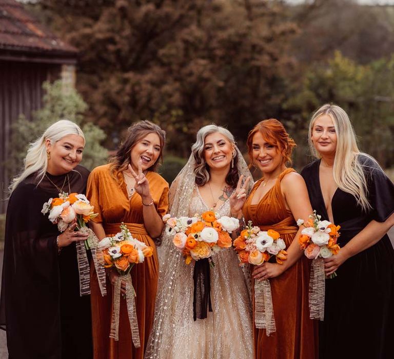 Bride in sleeveless deep v-neck sparkly gold wedding dress with sheer and gold polka dot wedding veil standing with bridesmaids in rust and black mismatched bridesmaid dresses