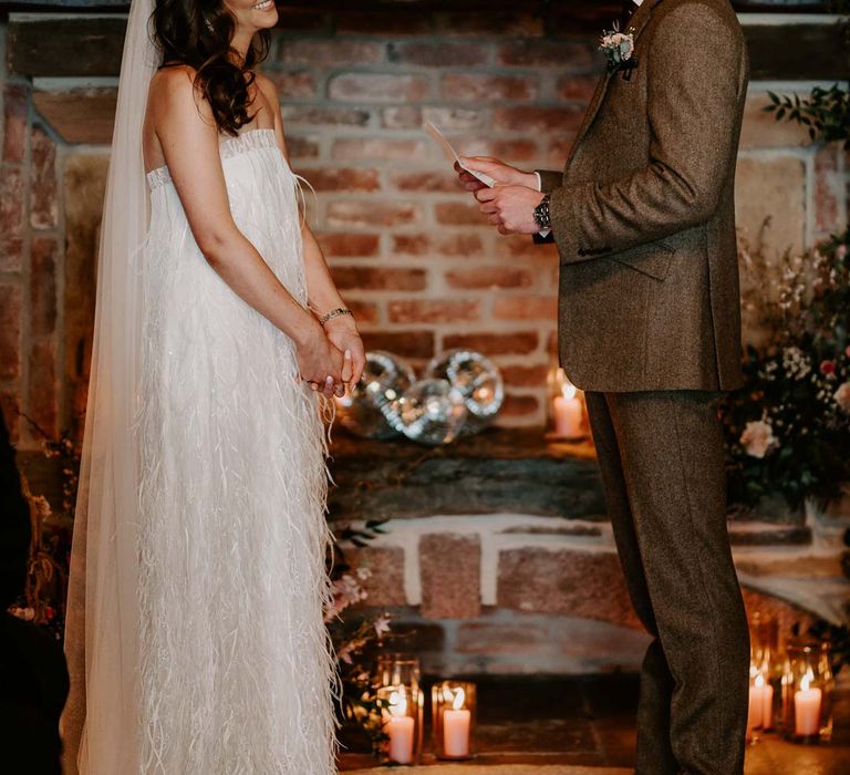 Groom in chocolate brown grooms suit doing wedding reading for bride in strapless ivory fringed crepe gown and chapel length veil standing by large fireplace and disco ball wedding decor