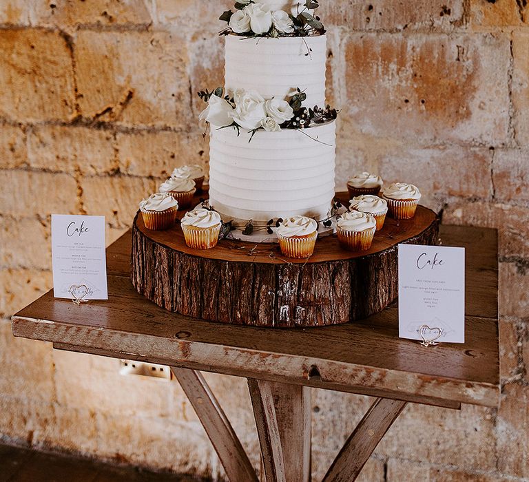 Three tier white frosted wedding cake with white rose wedding cake decorations for classic wedding surrounded by wedding cupcakes 