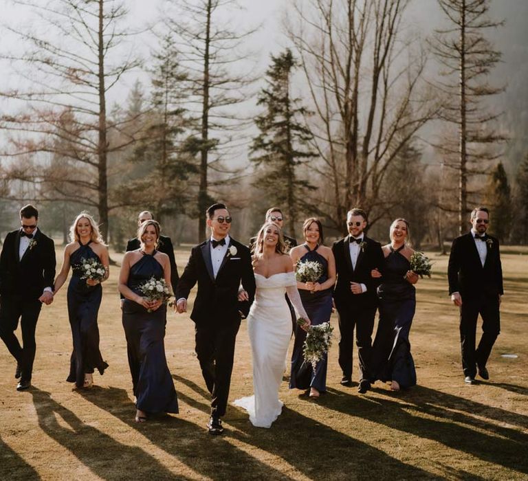 Groom in classic black tuxedo, black bowtie and white garden rose boutonniere walking with bride in off shoulder sparkly mermaid wedding dress and bridesmaids in satin halterneck navy bridesmaid dresses at French destination ski wedding